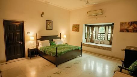 A wide shot of the heritage room at Mahendra Niwas, featuring a large, dark wooden bed with a vibrant green patterned duvet, and a cozy window seat.