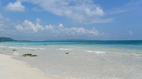 Tropical beach with white sand, turquoise water, and distant forest under a partly cloudy sky.