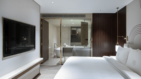 Bedroom with a plush bed facing a TV on the wall and a glass partition to the bathroom area at Merlis Hotel, Coimbatore