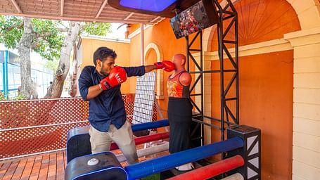 Man using a boxing arcade machine at MGM Dizzee World, wearing red gloves and punching a dummy in a shaded outdoor setup.