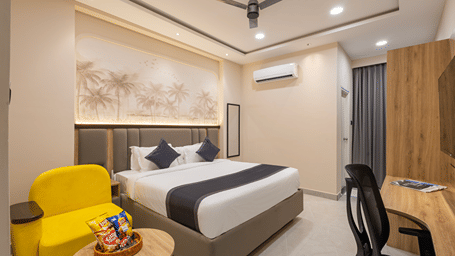 Premium room with a king bed, grey headboard, sepia palm mural, yellow armchair, snack basket and work desk at Monday Hotels & Suites Madhapur, Hyderabad.