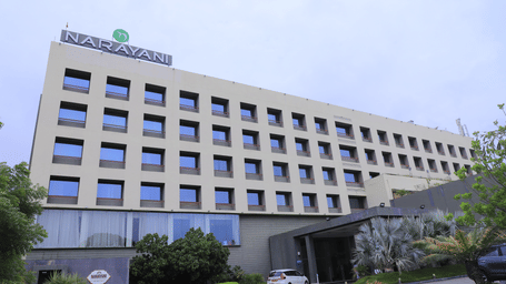 Close-up shot of the facade view of Narayani Heights Hotel & Resorts, Ahmedabad with trees and parking space.