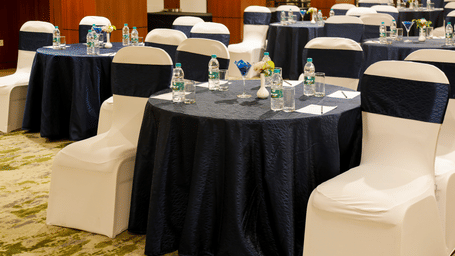 Corporate event setup featuring round tables with dark blue cloths, white chair covers with navy sashes, and bottled water in a conference hall.