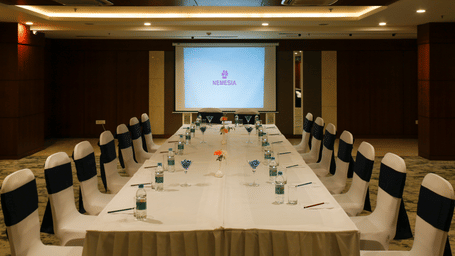 Long boardroom table set up for a corporate meeting, with chairs featuring navy sashes facing a large projector screen at the end of the room.