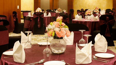 A round table covered in a satin table cloth with small white plates, glasses, and a glass flower vase with flowers.