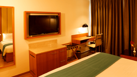 Modern hotel bedroom interior showing a flat-screen television, a wooden study desk with a chair, a large wardrobe mirror, and a green bed runner.