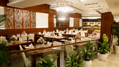 Spacious hotel restaurant dining area featuring wooden wall panelling, set tables, potted plants, and modern chandelier lighting.