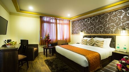 A  room at New Orchid Lords Inn Gangtok includes a comfortable bed with decorative pillows, a bedside table with water bottles, a functional desk with a chair, and a mounted TV for entertainment.