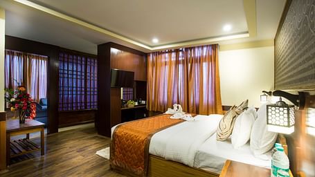 A room at New Orchid Lords Inn Gangtok contains a bed with pillows and folded towels, bedside tables with lamps and water, curtains, and a table with flowers.