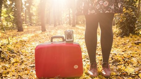 A close up shot of a woman standing next to a red suitcase in a forest