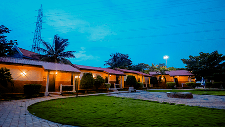 Exterior view of cottages with red roofs and green lawns at Nirantara Resort & Spa, Bengaluru, at dusk with lights illuminating the pathways.