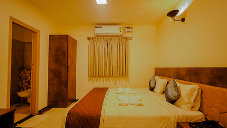 Side angle view of a room at Niratara Resort and Spa featuring a bed, window with short curtains, an almirah, and the bathroom door.