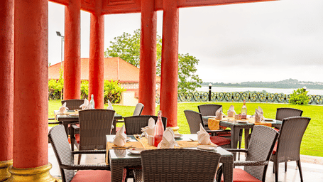 The outdoor dining area of the Marble Arch multi-cuisine restaurant in Bhopal at the Noor-Us-Sabah Palace overlooking the lush green lawn and the lake under a cloudy sky in the background.