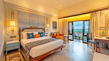 The brightly lit interiors of the Premium Club Room at Noor-Us-Sabah Palace, Bhopal, showing the bed's patterned headboard, bedside lamps, and a dedicated workspace, overlooking the beautiful views from the balcony.