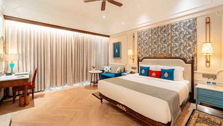 A wide-angle shot of the Royal Club Room at Noor-Us-Sabah Palace, Bhopal, featuring a work desk in the corenr, a plush double bed in a brightly lit room.