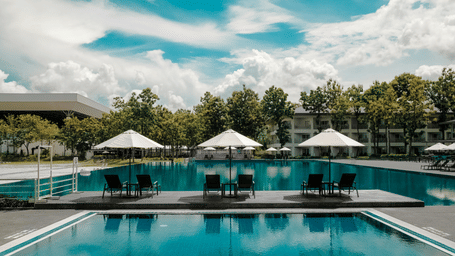 A wide view of a large, clear blue swimming pool surrounded by palm trees and curved patio seating areas under a bright sky at Ocean Sprays.