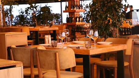 A warmly lit restaurant interior features set tables with wine glasses and chairs, with wine racks and greenery in the background.