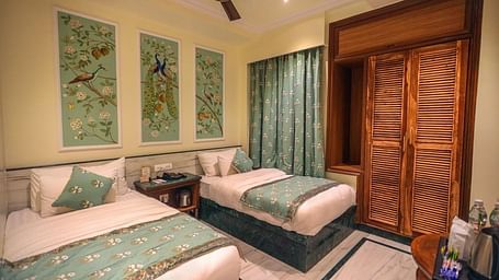 Diagonal view of the spacious room with twin beds placed side by side, a wooden bedside table in the middle, three photo frames above the headboard, and wooden storage | Panna Vilas Udaipur