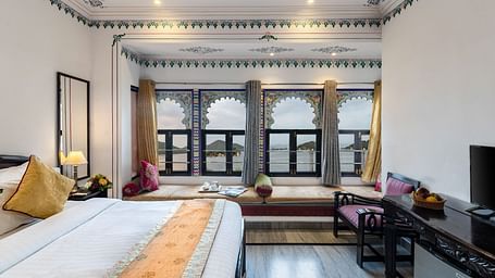 A side view of the bedroom featuring a spacious bed, a wooden table under the wall-mounted television, and bay window seating with curtains pulled to the side at Panna Vilas Palace