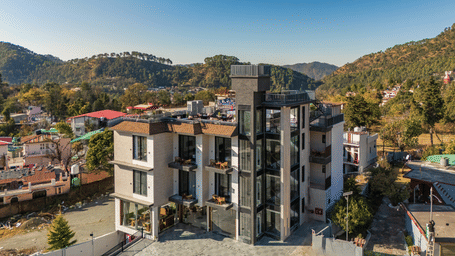 A high-angle shot of a contemporary 4-storey building surrounded by hills and local residential structures | Perfectstayz Premium Kapilaz, Bhimtal