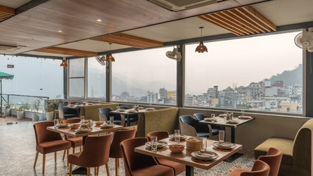 Outdoor rooftop restaurant with multiple wooden tables and chairs set for dining, overlooking a cityscape with hills in the background under clear skies.