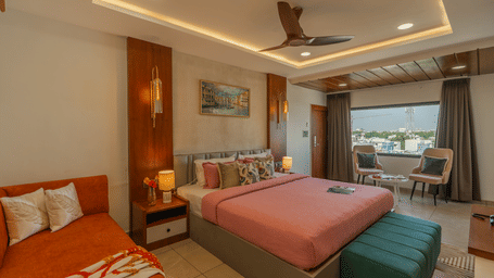 A large suite at Perfectstayz Premium Shree Narayana, Ujjain featuring a bed with pink linens, an orange sofa, and a seating area by the window.