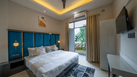 A spacious bedroom featuring a bed with a teal tufted headboard, large window with sheer curtains, a desk, a ceiling fan, and a television - Perfectstayz Value Aalaya Suites, Haridwar