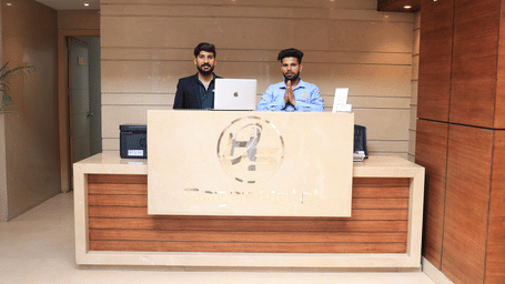A hotel reception desk made of wood and light stone, with 2 male staff members standing behind it | Perfectstayz Value Sagar