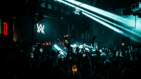 Audience members recording a live electronic music performance as bright laser beams illuminate the packed indoor venue.