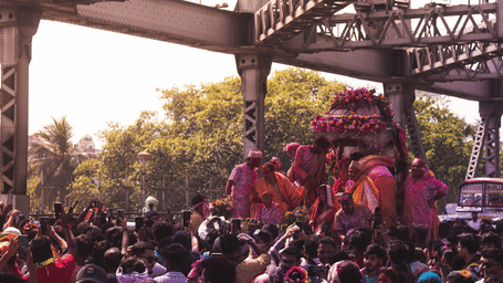 Dense crowd of devotees with phones raised, carrying a flower-decorated palanquin and idol beneath the steel truss girders of a large iron bridge.