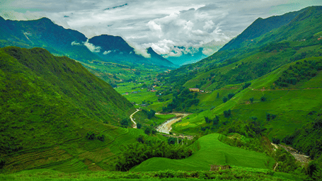 Panoramic view of a lush green mountain valley with terraced fields, a winding river, and clouds hovering over surrounding peaks.