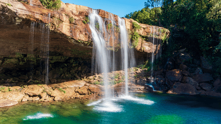 The scene features a wide, tiered waterfall cascading from a rocky, flat-topped cliff into a vibrant turquoise-blue emerald pool below.