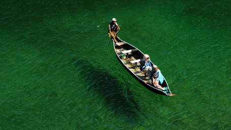Wooden canoe with three rowers gliding across clear emerald-green river water, the boat casting a long shadow on the riverbed.