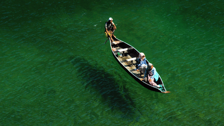 Aerial view of a traditional wooden Khasi boat with 3 people crossing clear emerald-green river waters near Dawki in Meghalaya.