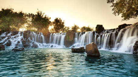 Wide curtain of waterfalls flowing over rocks into a turquoise pool at sunset with large boulders in the foreground and warm orange sky