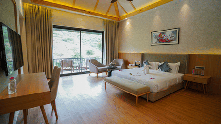 A modern hotel room with wooden floors and a large bed covered in rose petals, a desk, and a large window overlooking a balcony.