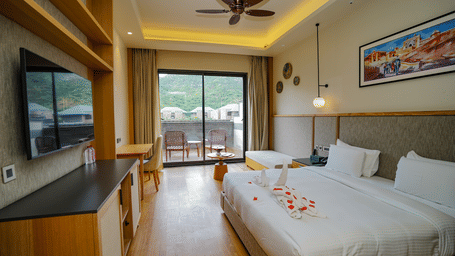 A well-lit hotel room with a large bed, a television, and a sliding glass door leading to a patio. There are rose petals on the bed.