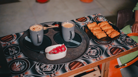 Two mugs of hot coffee served with a plate of evening snacks on a patterned tablecloth.