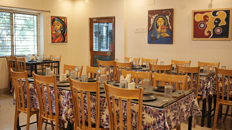 An organised restaurant seating area with wooden tables, chairs, and colourful paintings displayed on the wall.