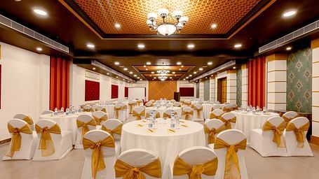A spacious banquet hall with round tables covered in white cloths and gold chair covers. The hall has a modern design with dark wood accents and a crystal chandelier.