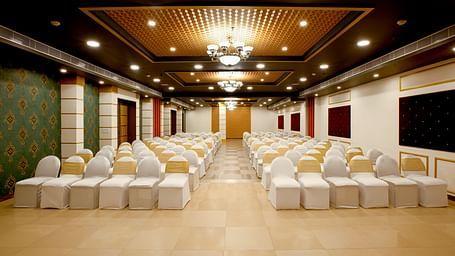 A spacious banquet hall with chairs covered in white cloths and gold chair covers. The hall has a modern design with dark wood accents and a crystal chandelier.