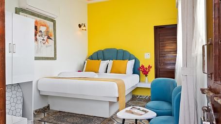 A cosy bedroom with a accent wall, a bed with white and mustard linens, and teal armchairs next to a small table.