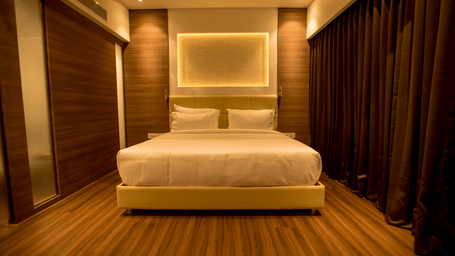 A modern king size bed in a hotel room is well-lit by soft backlighting against a dark headboard at S Hotels, Chennai.