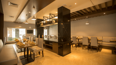 A dimly lit restaurant with tables with light coloured seating on the right at S Hotels, Chennai.