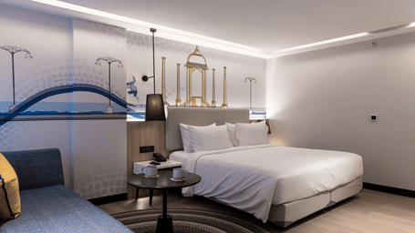 A Premium Room at Sands Point Hotel, Pondicherry, featuring a king bed in crisp white linen, a black reading lamp, a round dark table with a grey sofa.