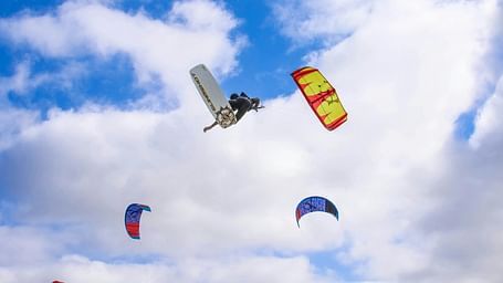 Multiple colourful kite surfers are airborne against a cloudy blue sky.