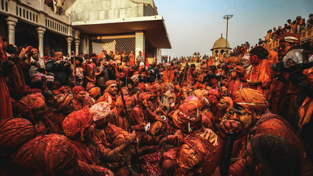 A large crowd celebrates the Holi festival, covered in vibrant orange and red powder near a traditional temple building.