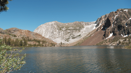 A serene panorama of a lake on the foothills of a mountain with surrounding snow-capped peaks under a clear blue sky.