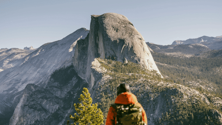 Back shot of a mountain climber wearing an orange jacket with a bag pack, looking at the panoramic Half Dome structure .