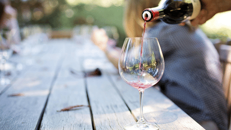 Red wine being poured into a large glass on a rustic wooden table during an outdoor lunch with friends.